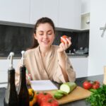 Portrait of beautiful smiling woman, writing her healthy menu, eating tomato while cooking, making
