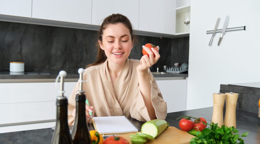 Portrait of beautiful smiling woman, writing her healthy menu, eating tomato while cooking, making